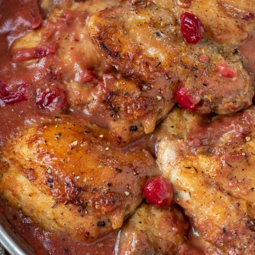 A skillet with golden brown pan seared chicken thighs in a cranberry gravy. There's whole cranberries in the sauce.