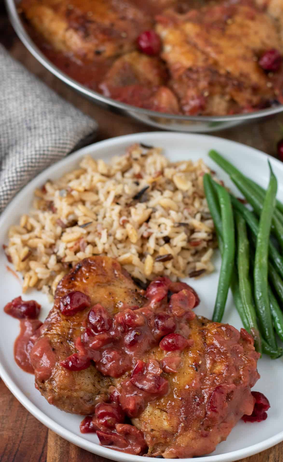 A white dinner plate with chicken thighs topped with cranberry Dijon gravy. There's also a side of green beans and wild rice on the plate. You can see the skillet with the rest of the chicken in gravy in the background.