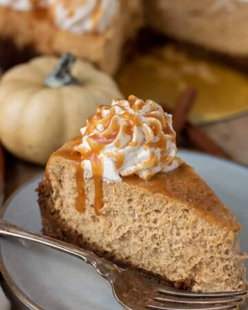 A small dessert dish topped with a slice of pumpkin cheesecake. It's got a dollop of whipped cream on top with a drizzle of salted caramel. There's a fork next to the slice of cake. In the background there's a small white pumpkin and the rest of the cheesecake