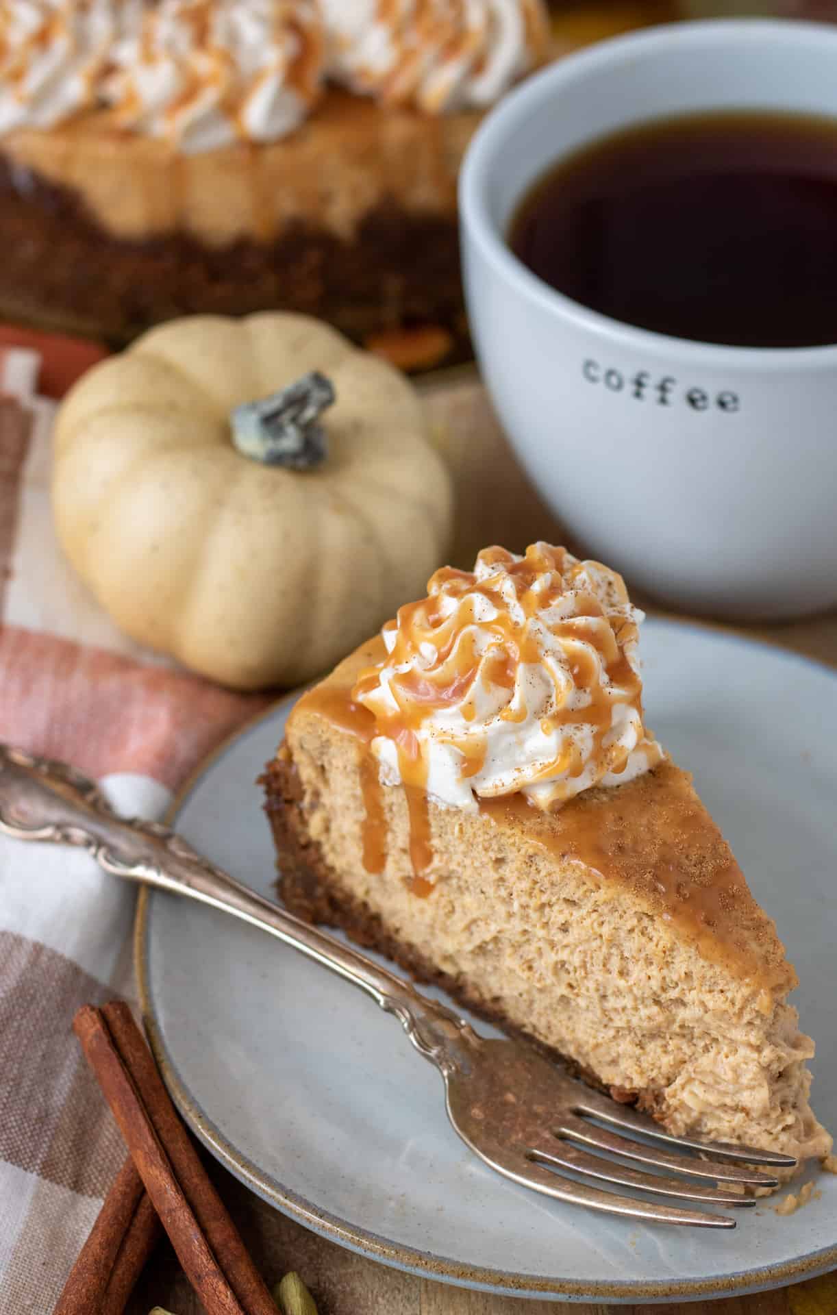 A small white dish with a slice of pumpkin cheesecake with gingersnap crust. There's a fork on the plate and the cheesecake has a dollop of whipped cream that's drizzled with salted caramel. There's a white mug of coffee in the background and a small white pumpkin sitting on an orange and white plaid dish towel.