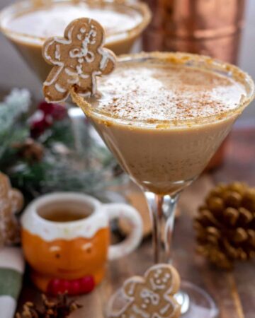 A festive Christmas gingerbread cocktail in a martini glass. It's got a gingerbread cookie placed on the rim and it's garnished with cinnamon. There's a cute little gingerbread espresso cup next to the martini glass with some pine cones and decorative tree branches with red berries.