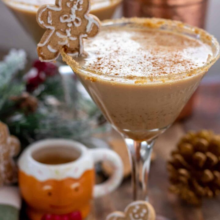 A festive Christmas gingerbread cocktail in a martini glass. It's got a gingerbread cookie placed on the rim and it's garnished with cinnamon. There's a cute little gingerbread espresso cup next to the martini glass with some pine cones and decorative tree branches with red berries.
