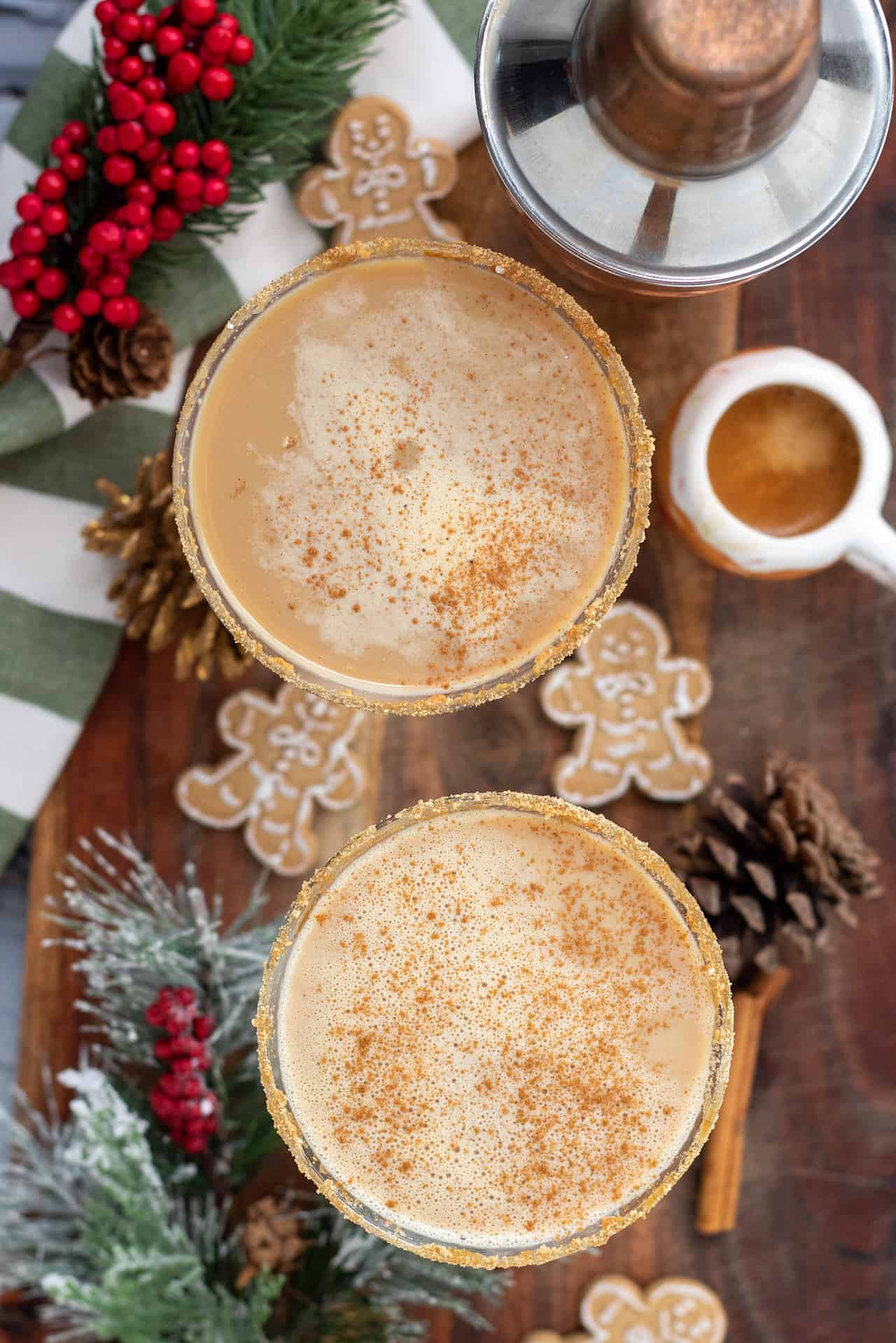An overhead shot of two martini glasses filled with gingerbread espresso cocktail. The martinis have a sprinkle of cinnamon on top. There's two gingerbread men cookies, pinecones, and a snow frosted tree branch in the background. There's a small cup of espresso too.