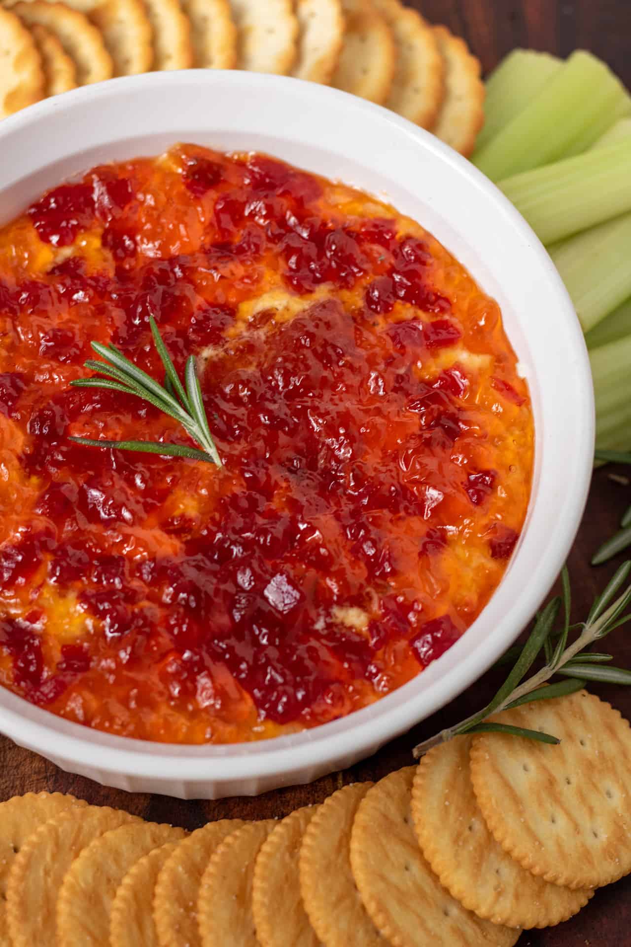 A round white serving bowl filled with cream cheese and pepper jelly dip. There's a sprig of fresh rosemary on top. You can see celery sticks and crackers next to the dip.