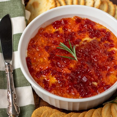 A round white serving dish filled with red pepper jelly dip. There's two kinds of assorted round crackers spread around it. There's a white and green striped kitchen towel next to the bowl with a knife spreader on top. There's a decorative pine berry tree branch in the background.