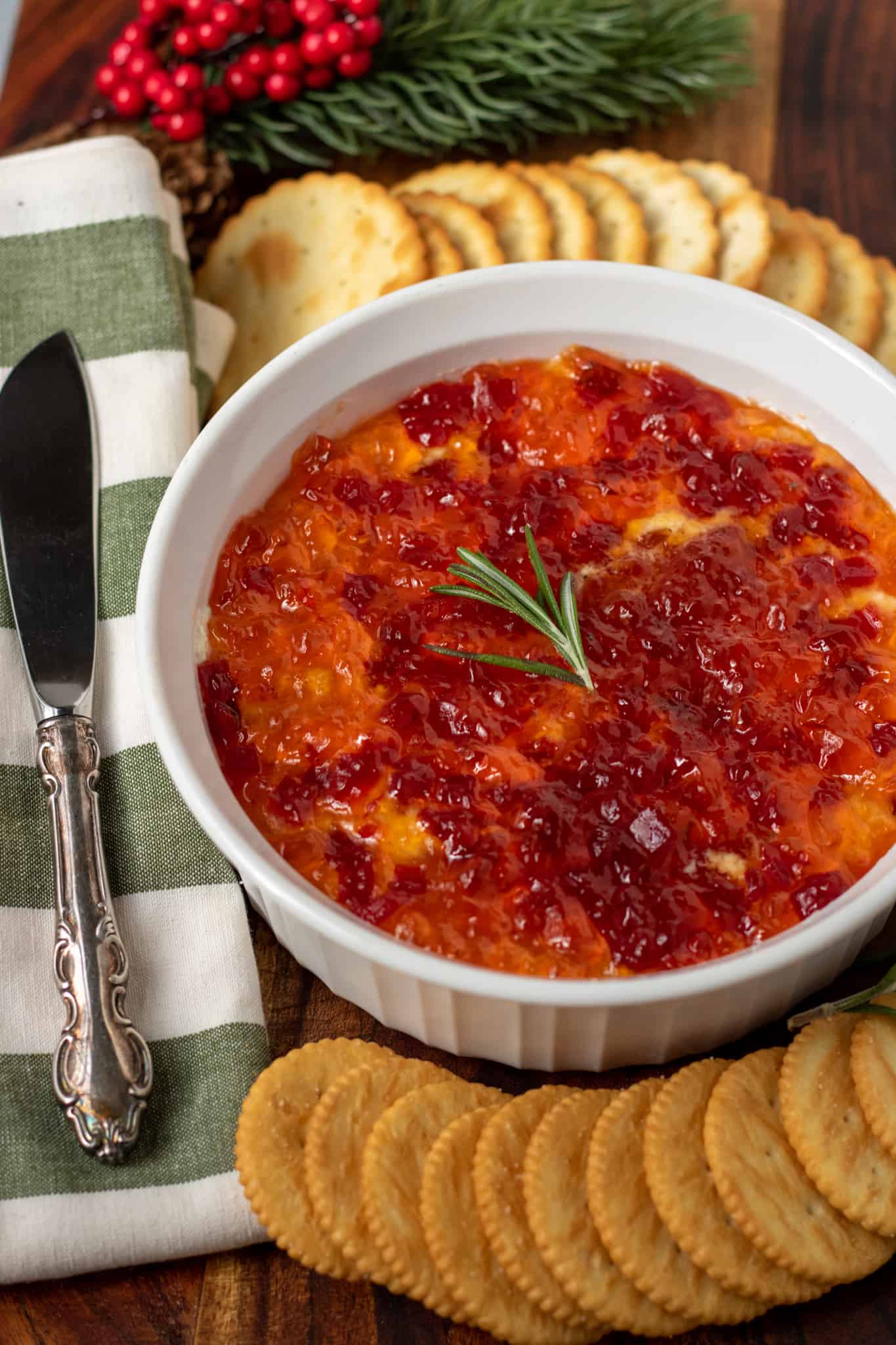 A round white serving dish filled with red pepper jelly dip. There's two kinds of assorted round crackers spread around it. There's a white and green striped kitchen towel next to the bowl with a knife spreader on top. There's a decorative pine berry tree branch in the background.