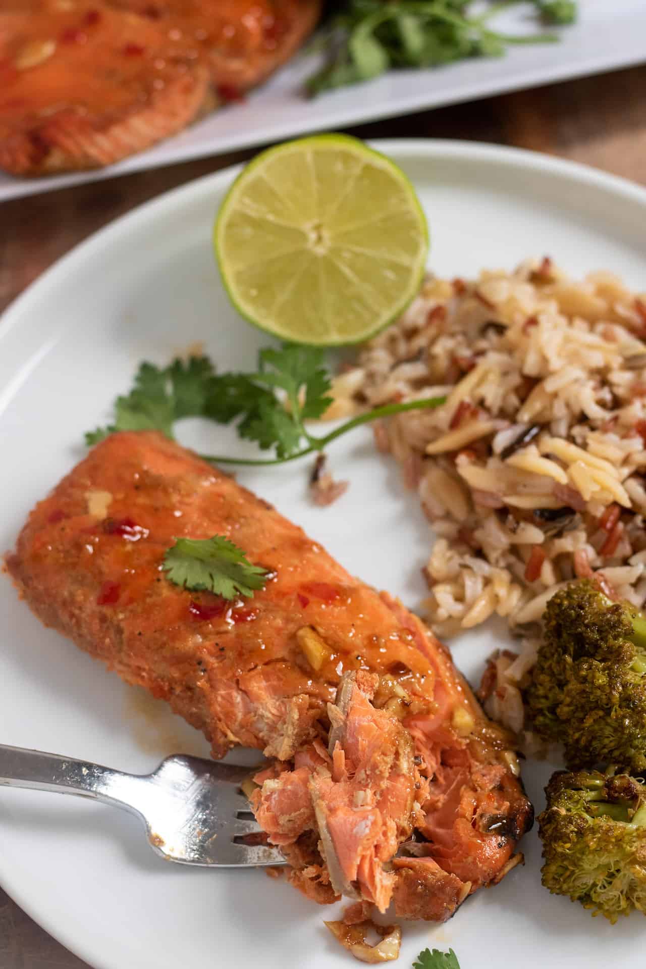 A round white dinner plate topped with a piece of salmon.  The salmon has a fork taking a piece from it.  There's wild rice and broccoli next to the salmon on the plate with a half of a lime and cilantro.