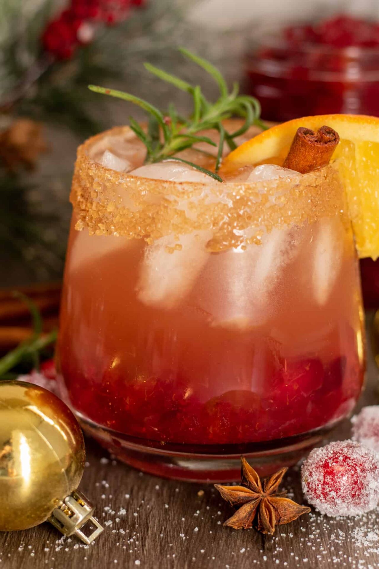 A short cocktail glass filled with holiday cranberry bourbon cider.  It's garnished with a sprig of rosemary, orange slice and cinnamon stick.  It has a cinnamon sugar rim.  There's a gold ornament next to the glass with sugared cranberries and whole cinnamon sticks.