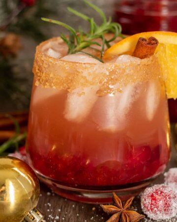 A short cocktail glass filled with holiday cranberry bourbon cider. It's garnished with a sprig of rosemary, orange slice and cinnamon stick. It has a cinnamon sugar rim. There's a gold ornament next to the glass with sugared cranberries and whole cinnamon sticks.