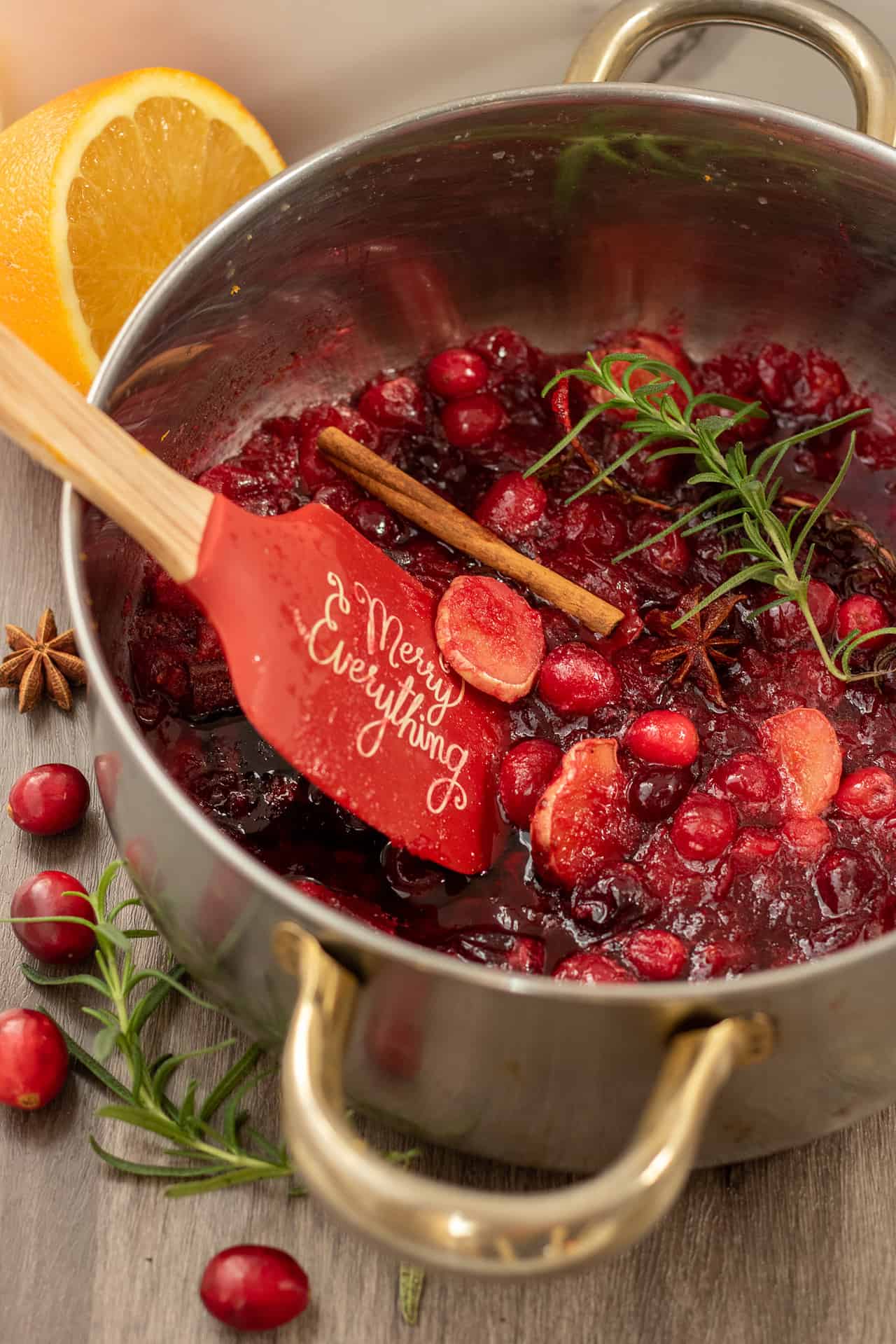 A silver sauce pan with gold handles filled with fresh cranberry syrup with whole cinnamon sticks, star anise, rosemary sprig, and ginger pieces.