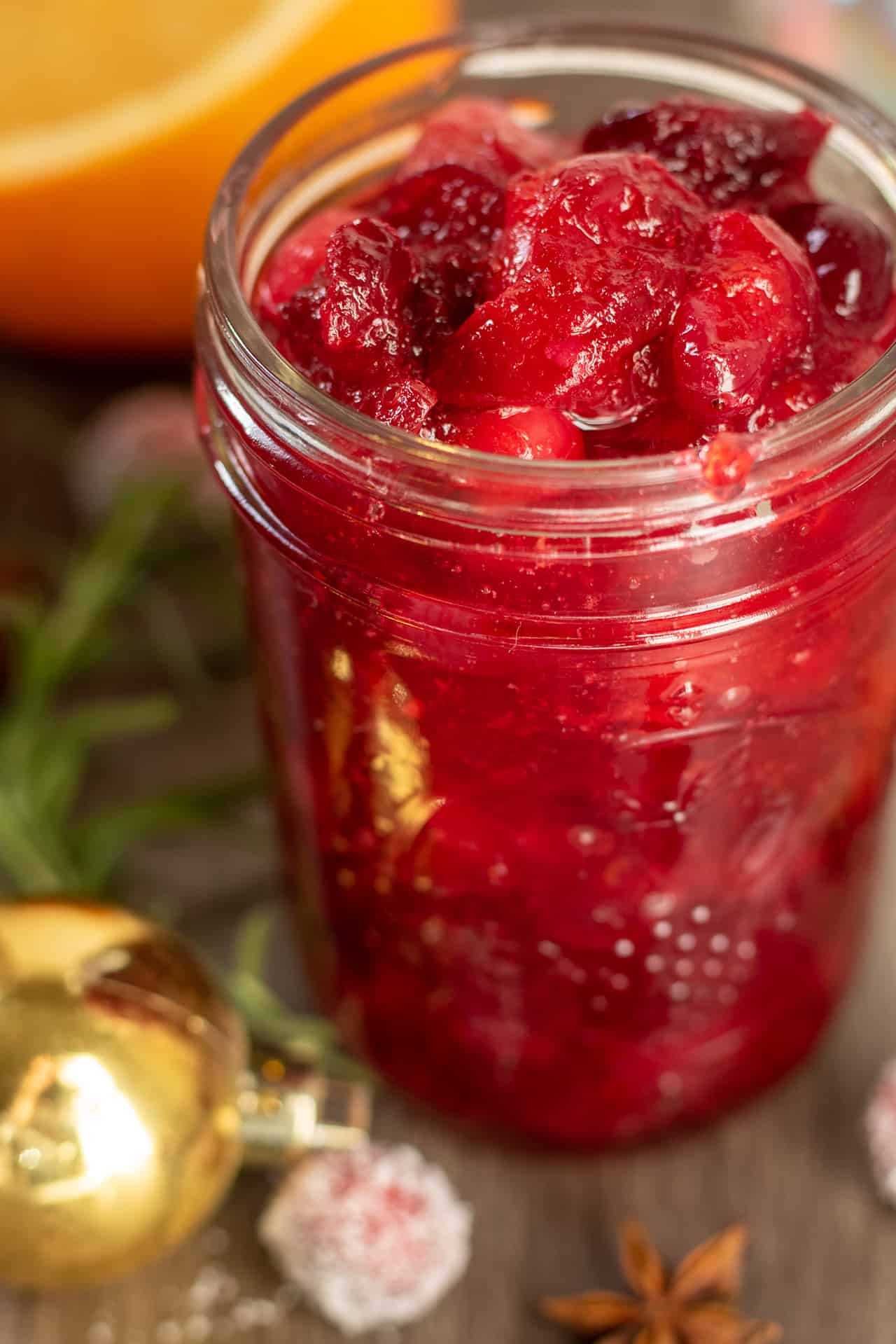 A mason jar filled with cranberry spiced syrup for a holiday cocktail. There's a gold ornament next to the jar with a sprig of rosemary and fresh cranberries