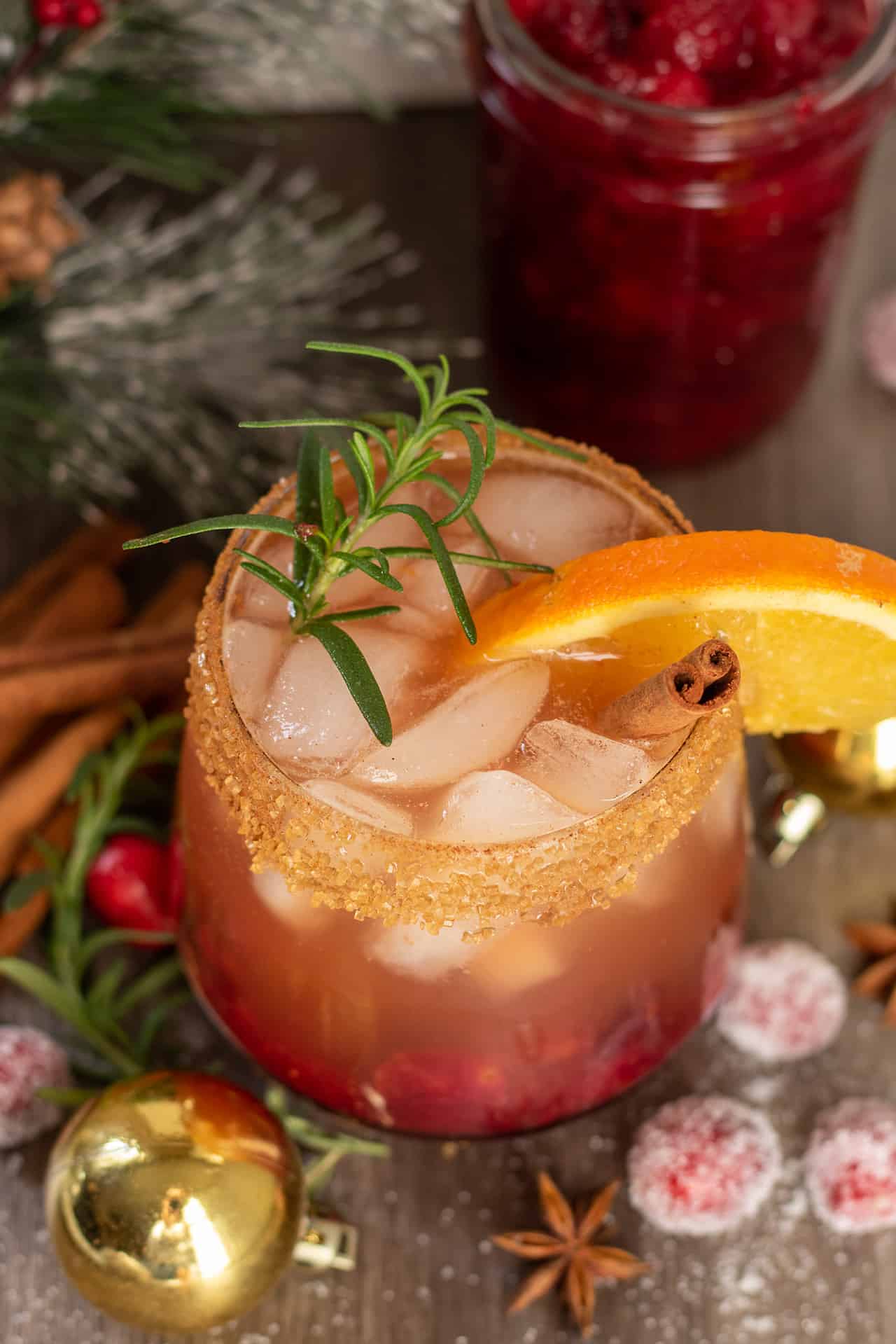 A cocktail glass filled with ice and cranberry bourbon cider. It's got a orange piece hanging on the side of the glass with a cinnamon stick and sprig of fresh rosemary. You can see sugared cranberries next to the glass with cinnamon sticks and a jar of cranberry syrup in the background