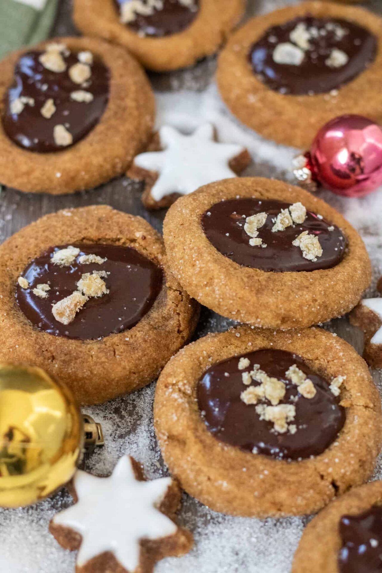 Gingerbread thumbprint cookies that are filled with chocolate ganache and topped with candied ginger pieces.  There's a small gold ornament and sugar in the background that looks like snow.