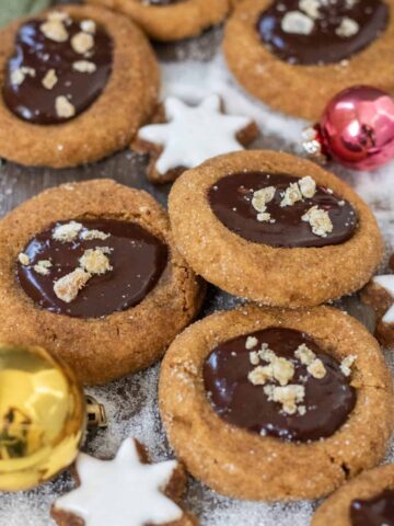 Gingerbread thumbprint cookies that are filled with chocolate ganache and topped with candied ginger pieces. There's a small gold ornament and sugar in the background that looks like snow.