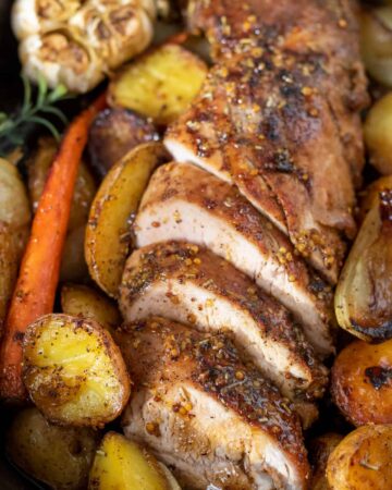 A sliced pork tenderloin that's been baked in the oven. It's got roasted vegetables in the cast iron skillet with it. There's a head of garlic and fresh rosemary sprigs. The pork tenderloin is brown on the outside and juicy on the inside.