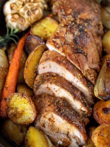 A sliced pork tenderloin that's been baked in the oven. It's got roasted vegetables in the cast iron skillet with it. There's a head of garlic and fresh rosemary sprigs. The pork tenderloin is brown on the outside and juicy on the inside.