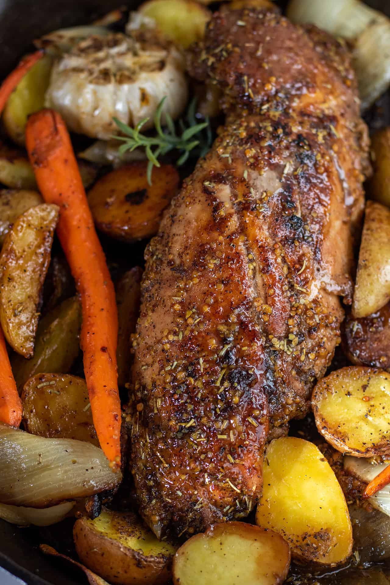 A whole oven roasted pork tenderloin that's been cooked in the oven. It's golden brown with a honey mustard glaze on top. There's roasted potatoes, carrots and onions around it.