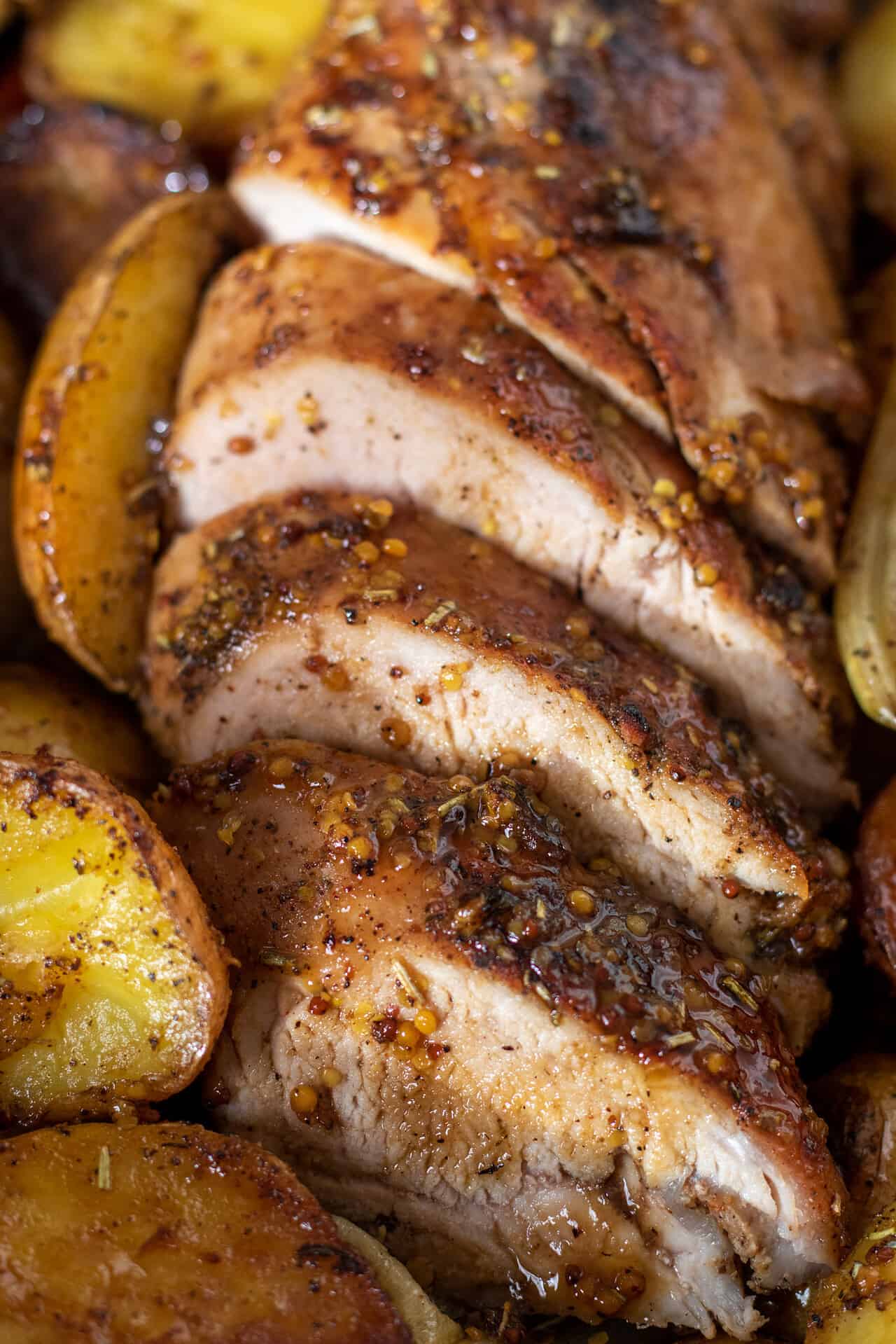A pork tenderloin that's been sliced for serving. There's a honey mustard glaze on top of it with roasted potatoes and carrots around the pan.
