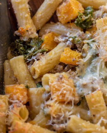 Rigatoni pasta with butternut squash cubes, kale, and ground sausage. There's cheeses on the pasta and melted mozzarella on top.