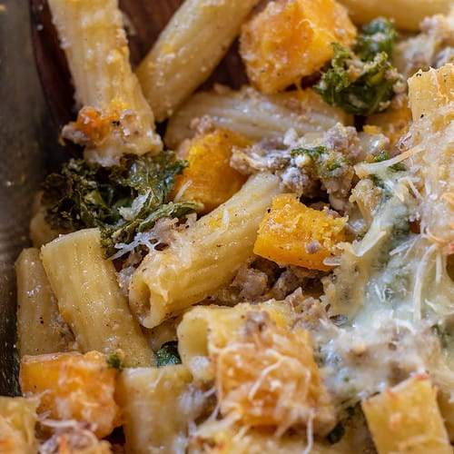 Rigatoni pasta with butternut squash cubes, kale, and ground sausage. There's cheeses on the pasta and melted mozzarella on top.