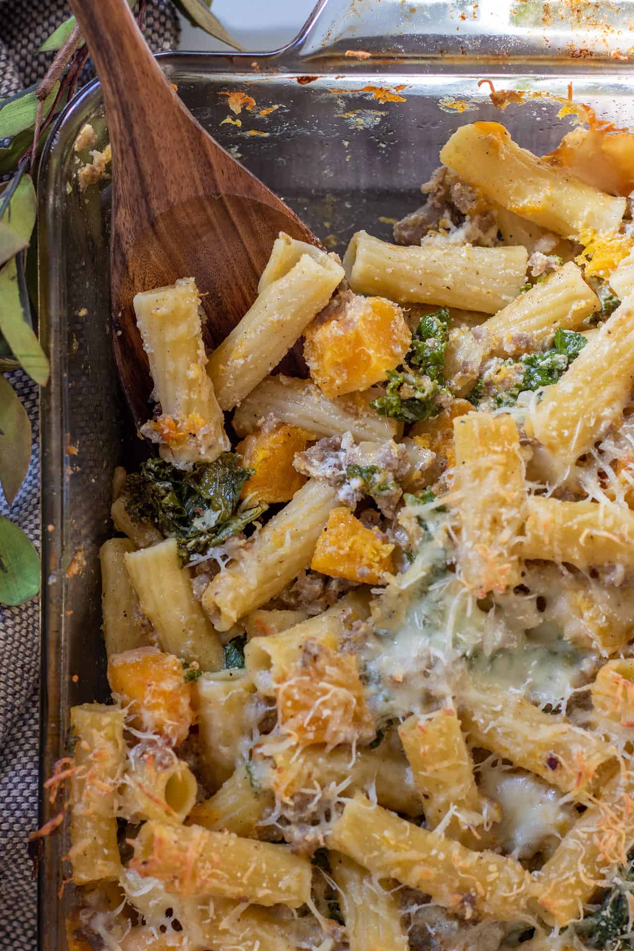 A glass baking dish filled with butternut squash pasta, sausage and kale. There's a wooden serving spoon taking a piece of the pasta.
