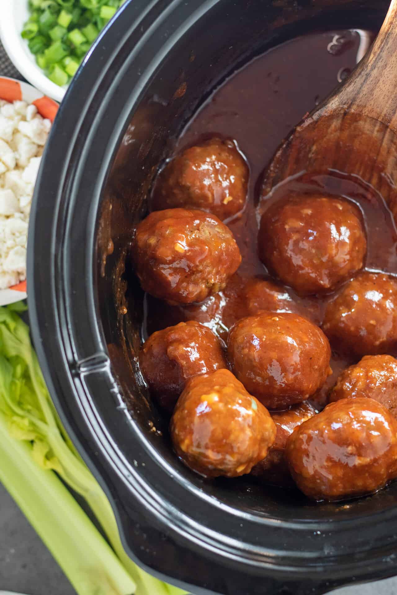 A black crockpot filled with buffalo bbq chicken meatballs. There's celery and blue cheese crumbles in the background