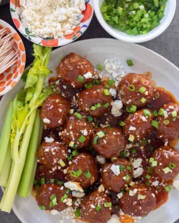 A round white plate with bbq buffalo meatball appetizers. There's topped with blue cheese crumbles and chopped scallions. There's 3 small bowls in the background, one with toothpicks, one with scallions and one with blue cheese. There's a couple of stalks of celery on the plate with the meatballs.