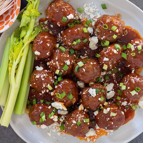 A round white plate with bbq buffalo meatball appetizers. There's topped with blue cheese crumbles and chopped scallions. There's 3 small bowls in the background, one with toothpicks, one with scallions and one with blue cheese. There's a couple of stalks of celery on the plate with the meatballs.
