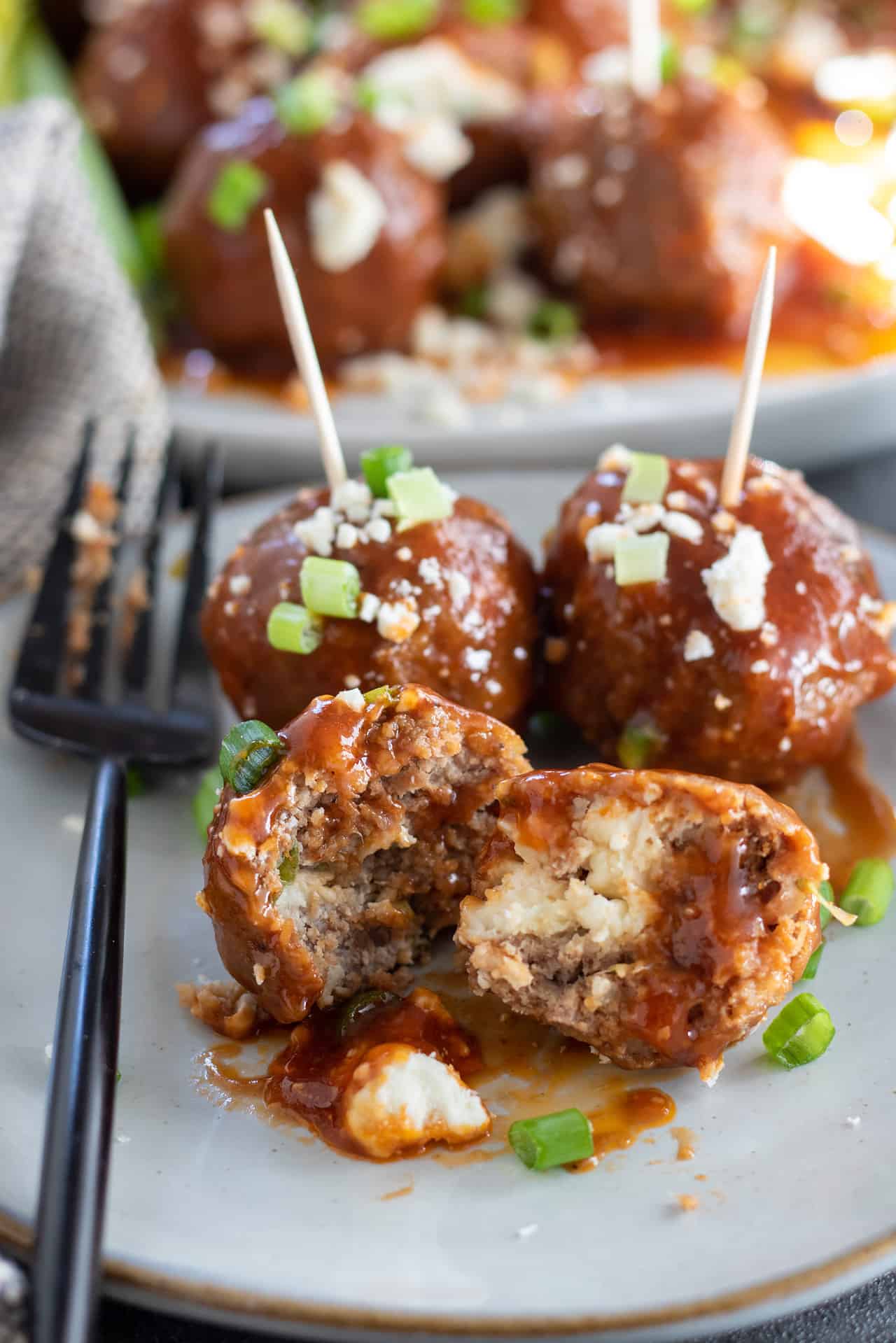 A small plate with 3 small meatballs on it. One meatball is cut open and it's stuffed with blue cheese. The other two meatballs have toothpicks in them for appetizers. There's a black fork on the plate and a bigger plate in the background with more buffalo chicken meatballs.