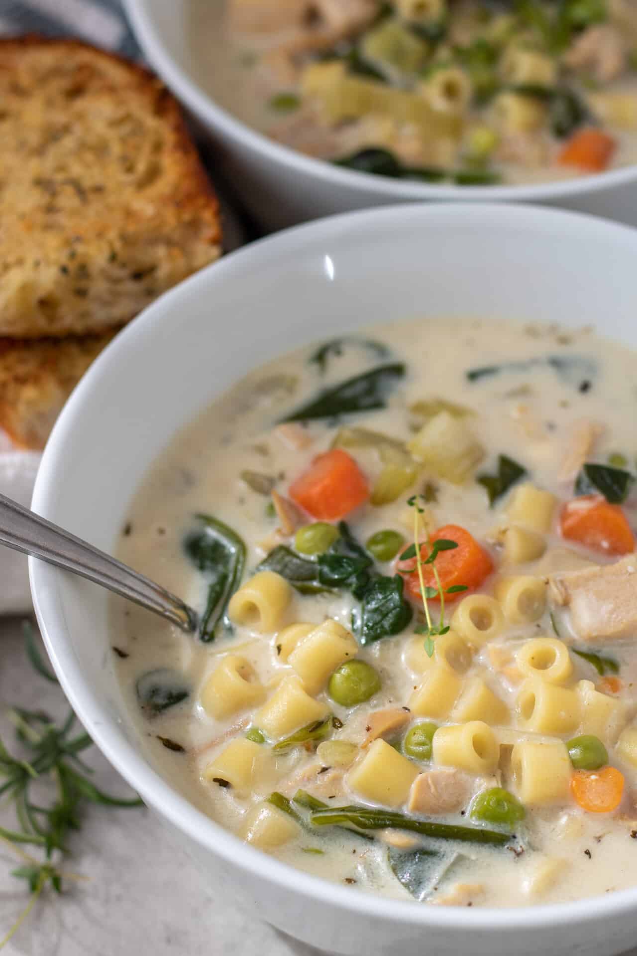 A white bowl with chicken soup with pasta, spinach, peas and carrots. The soup is creamy. There's two pieces of golden crusty garlic bread next to the bowl.