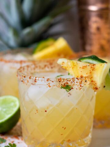 A spicy pineapple margarita. There's a slice of pineapple and lime on the edge of the glass and it has a spiced salted rim. There's half of a lime next to the glass and you can see the top of a cut pineapple in the background.