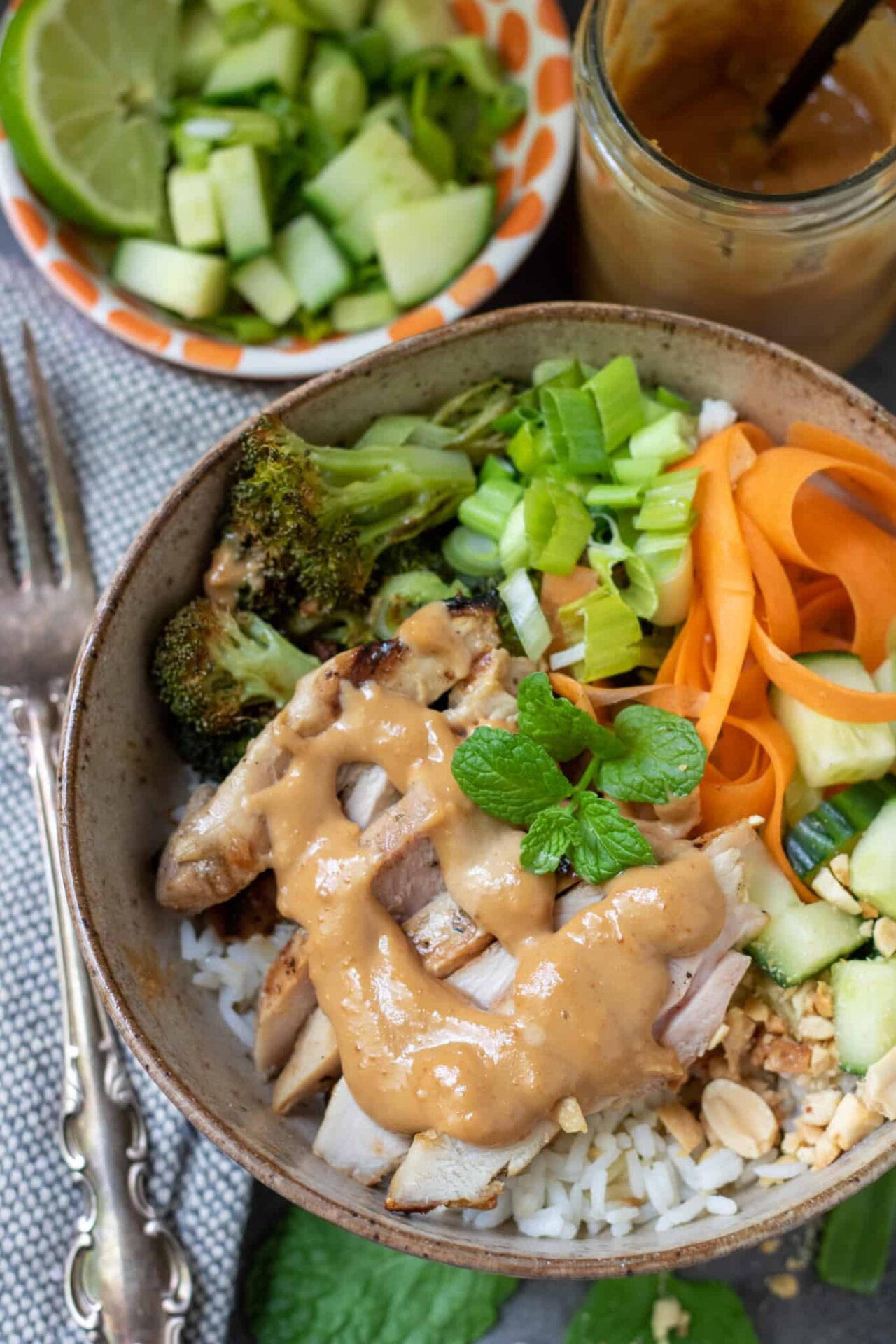 A bowl filled with rice, roasted broccoli, carrots and diced cucumber. There's sliced grilled chicken and a peanut sauce drizzled on top. There's a fork next to the bowl and a small bowl filled with diced cucumber in the background.
