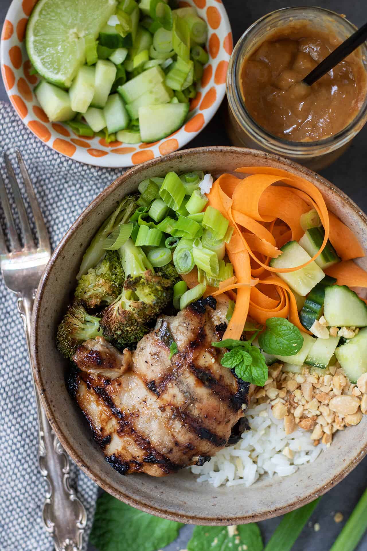 A Thai rice bowl with grilled chicken. There's crushed peanuts, cucumber, carrots, mint and broccoli on top of the bowl.