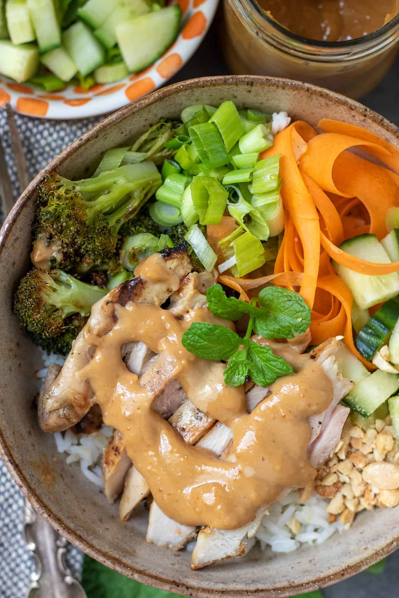 A close up of a Thai chicken rice bowl. There's a peanut sauce drizzled on top. The bowl has ribboned carrots, diced cucumbers, scallions and broccoli. There's a sprig of fresh mint on top of the rice bowl.