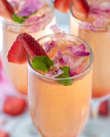 3 champagne flutes filled with sparkling rosé. They're topped with edible rose petals and a slice of fresh strawberry.