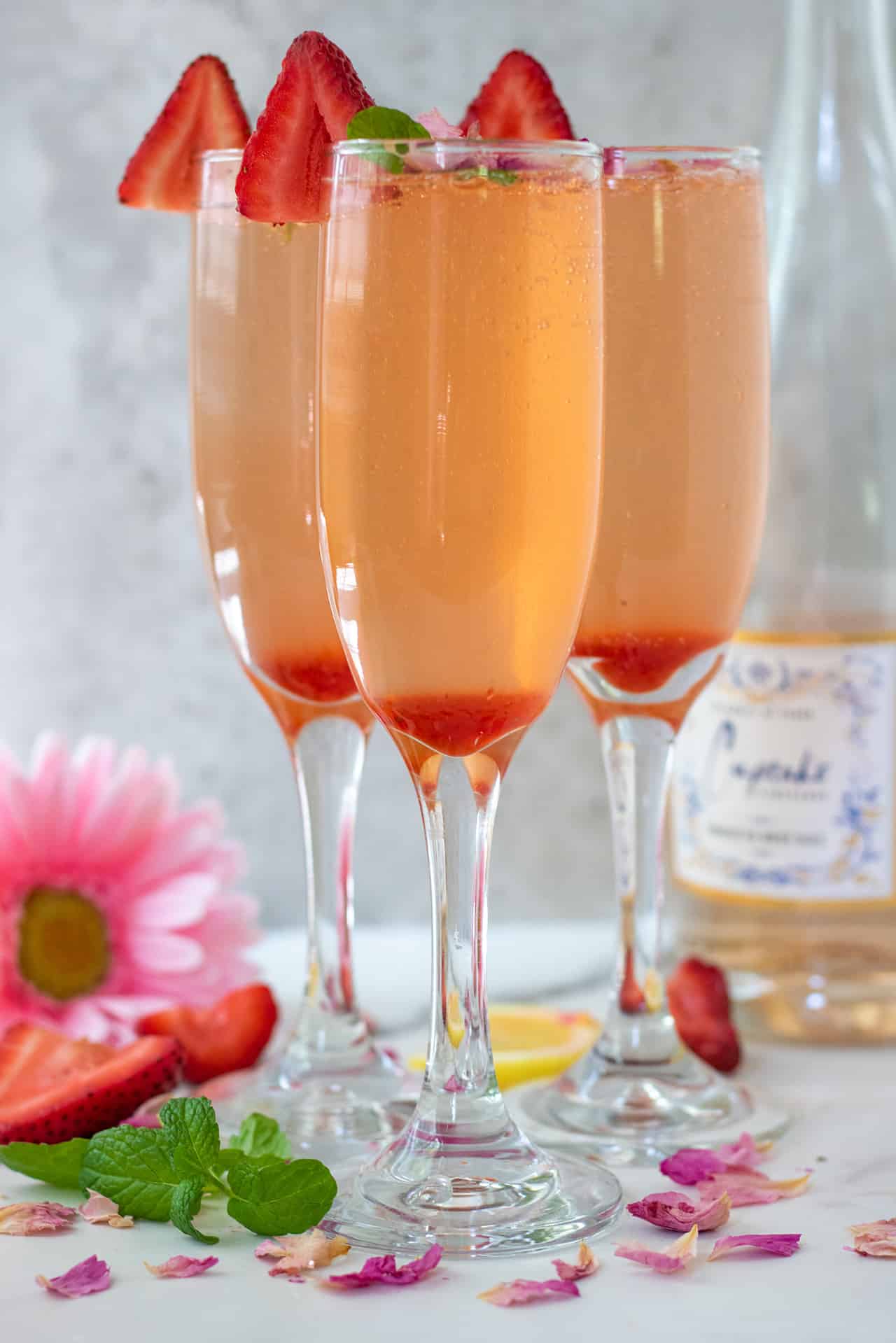 3 champagne flutes filled with strawberry mint syrup and topped with sparkling rosé champagne. There's a bottle of the rosé in the background with a pink daisy flower and fresh strawberries.