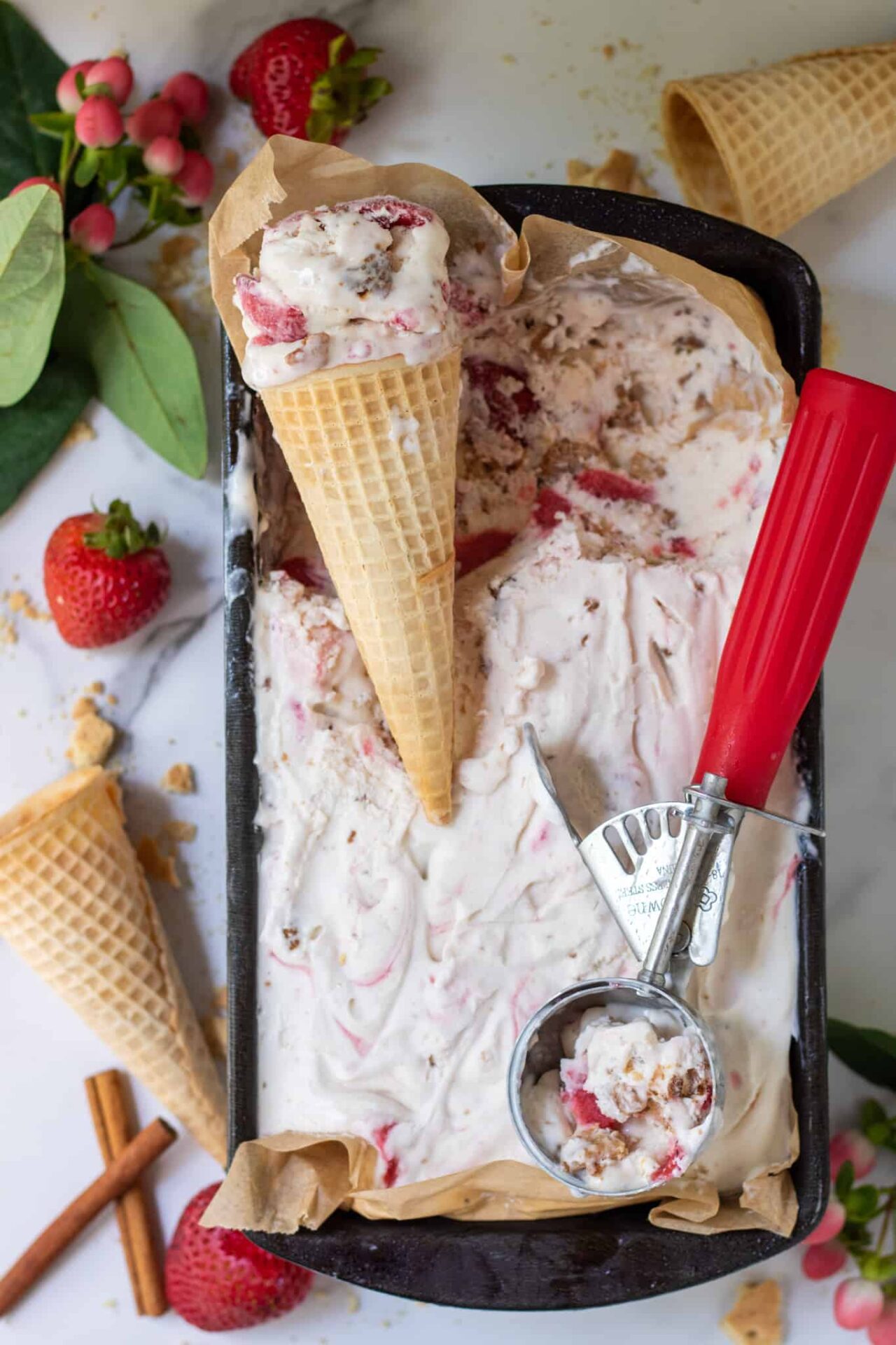 A loaf pan filled with strawberry ice cream.  There's a red ice cream scoop and two cones of ice cream.  There's whole strawberries in the background.