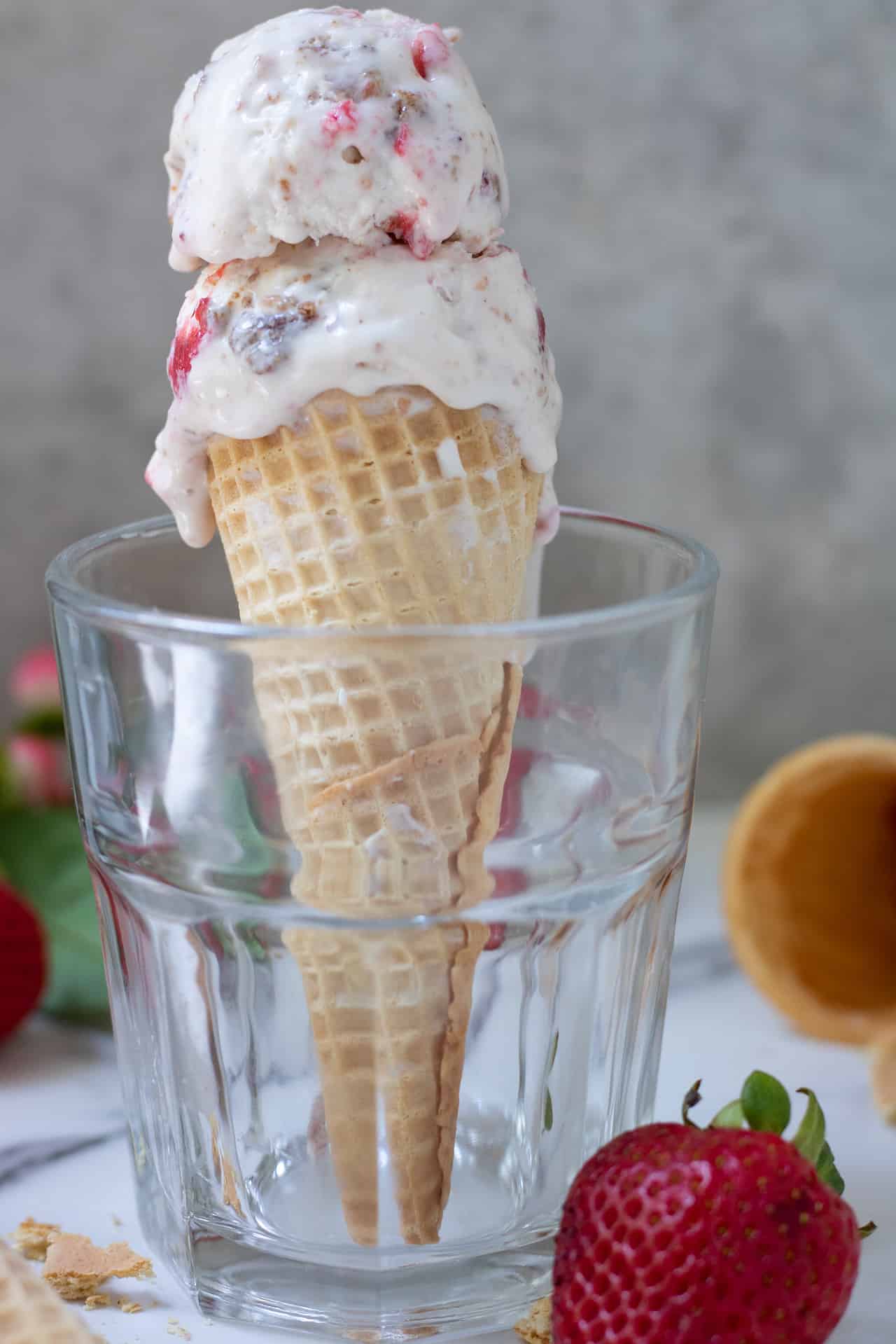 A short glass with an ice cream cone in it. There's ice cream dripping down the sides. you can see more ice cream cones and a strawberry in the background