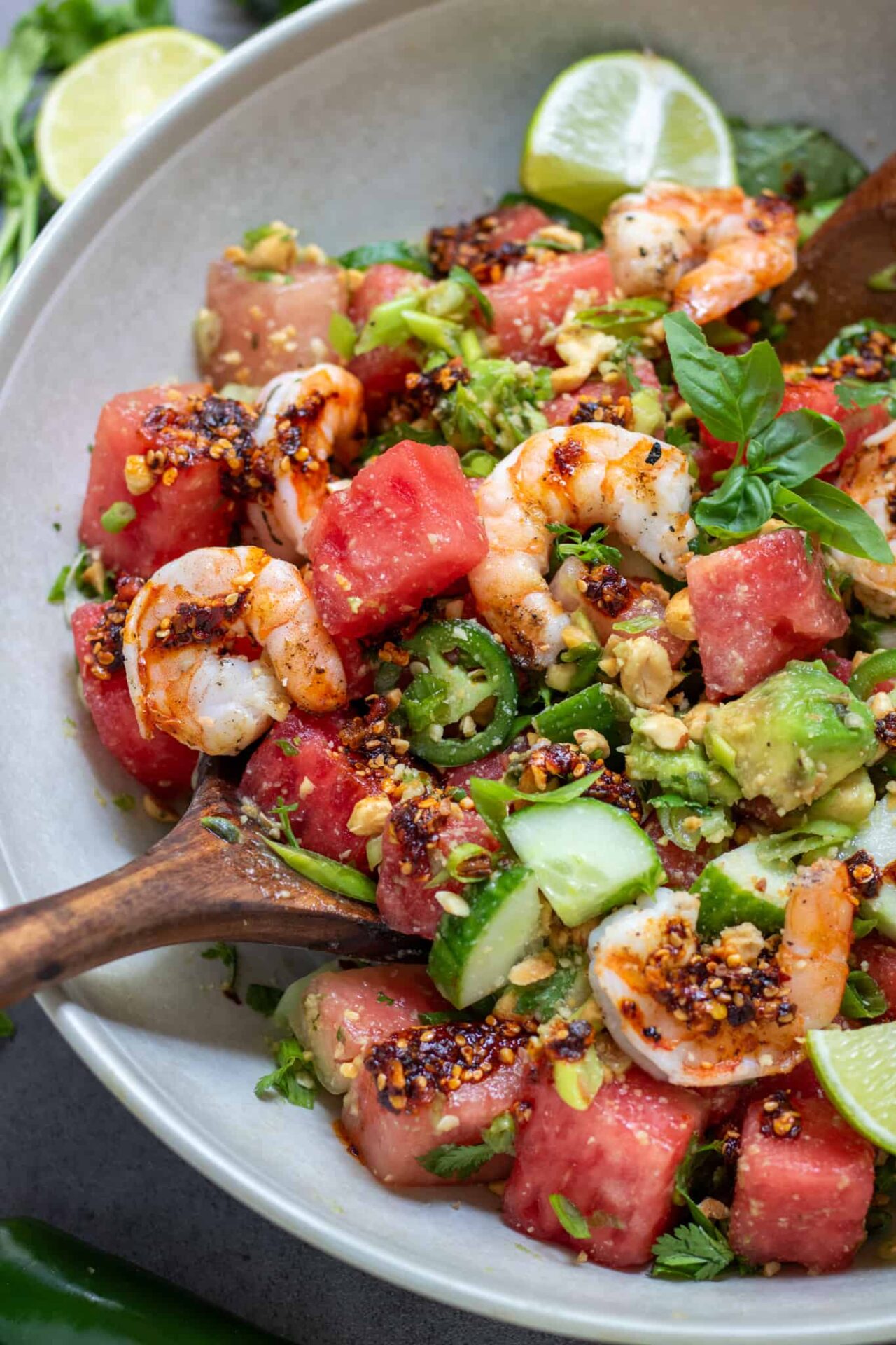 A large cream colored serving bowl filled with spicy watermelon salad. There's a wooden serving spoon in the salad. It's topped with chili crunch oil, and grilled shrimp.