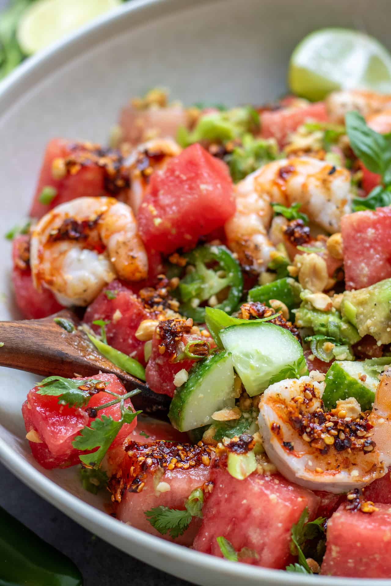 Watermelon salad with diced cucumbers, avocado and sliced jalapeño peppers. There's a wooden serving spoon in the salad bowl. It's drizzled with a chili crunch oil and topped with grilled shrimp.
