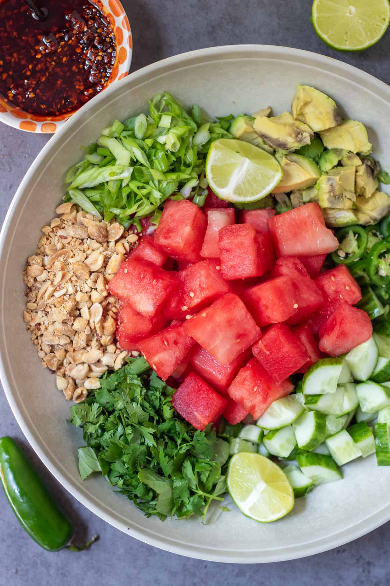 A large round serving bowl with cubes of watermelon, diced cucumbers, diced avocado, sliced jalapeños, sliced scallions, crushed peanuts, and fresh chopped herbs. There's a small bowl filled with chili crunch oil in the background.