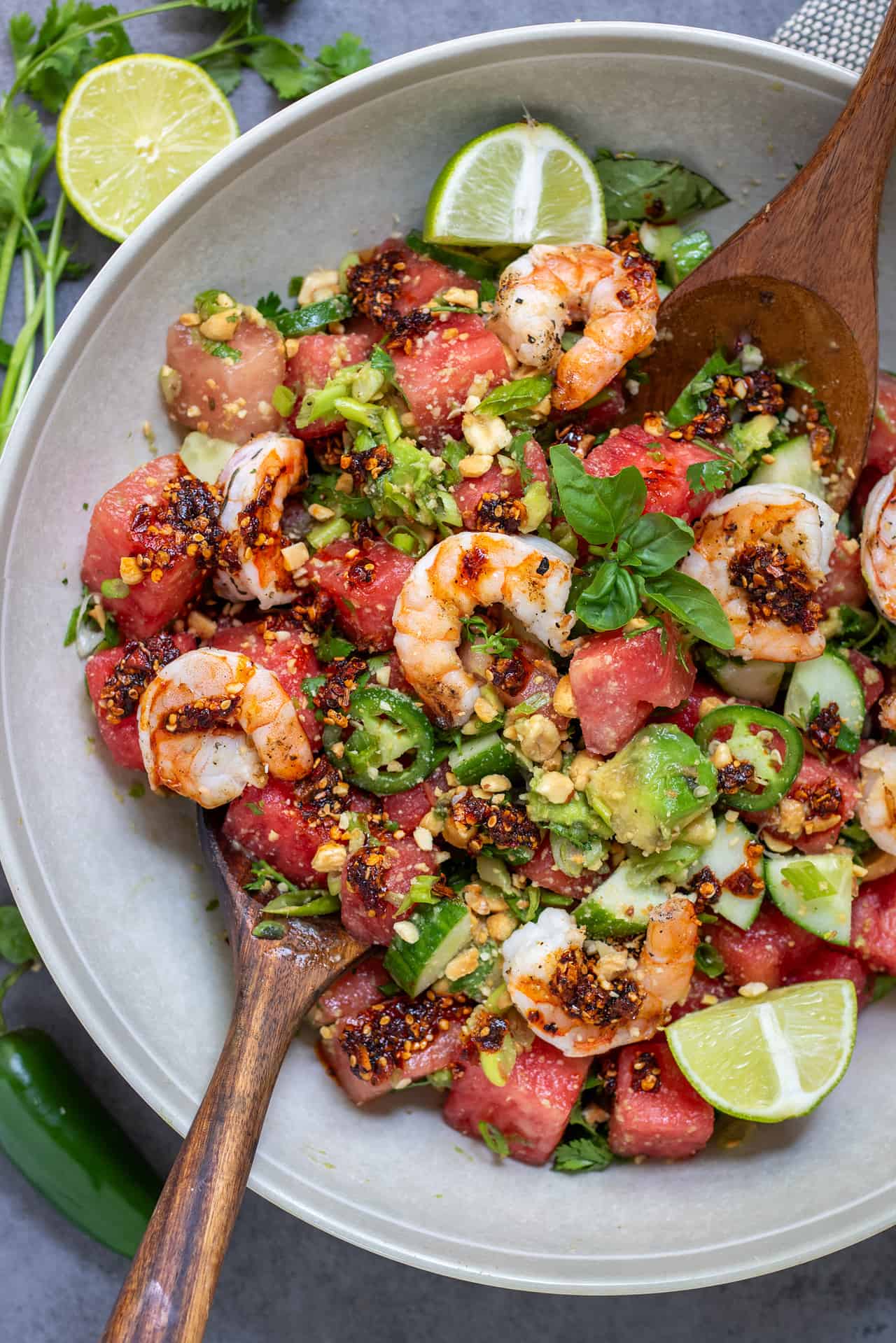 A large round salad bowl filled with spicy watermelon salad topped with grilled shrimp. There's a fresh sprig of mint in the middle and the salad is drizzled with chili crunch oil. You can see the diced cucumbers and avocado. Two serving spoons are in the salad bowl.