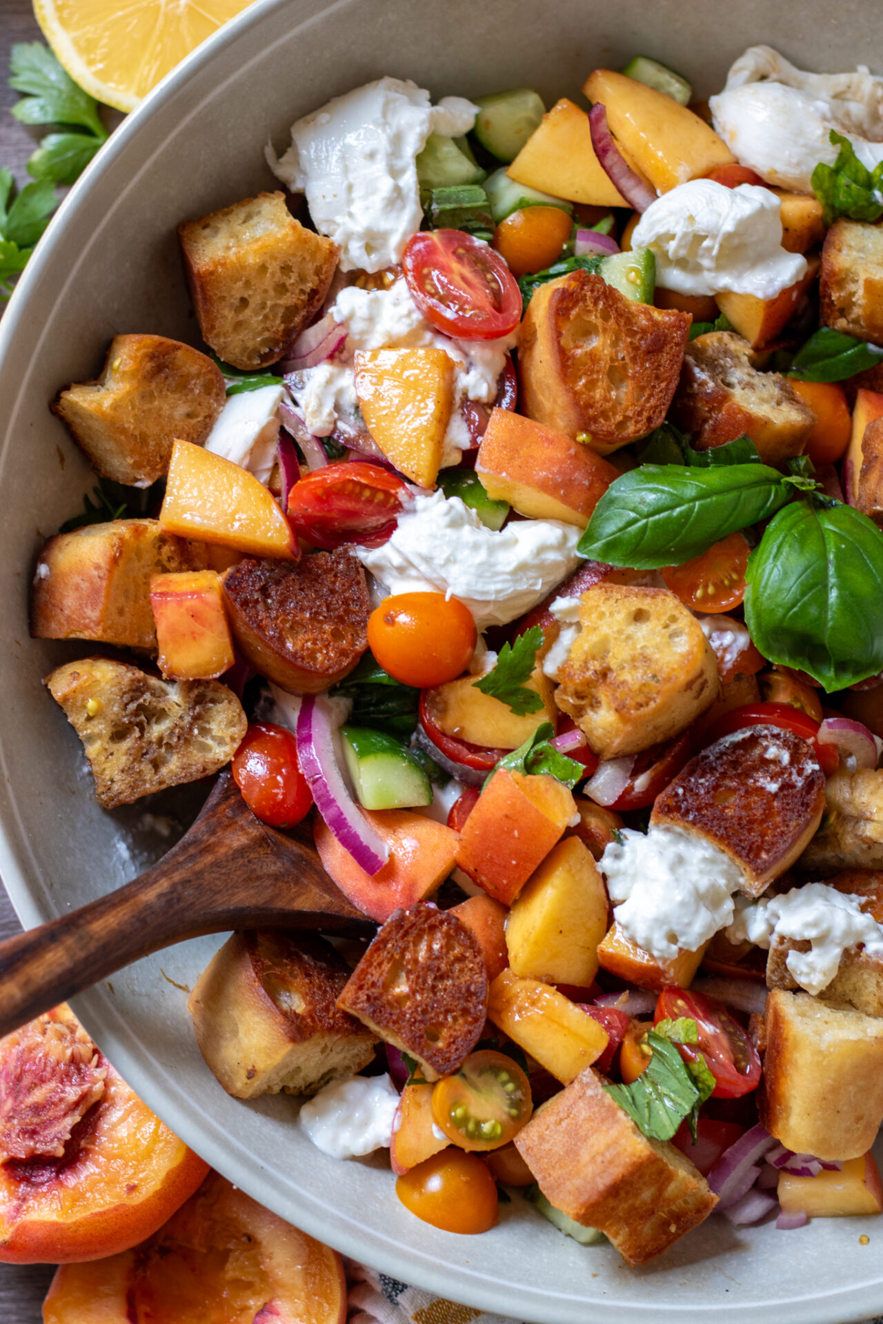 Peach panzanella salad with burrata with tomatoes, cucumbers and red onion. It's got a wooden serving spoon dug into the bowl.