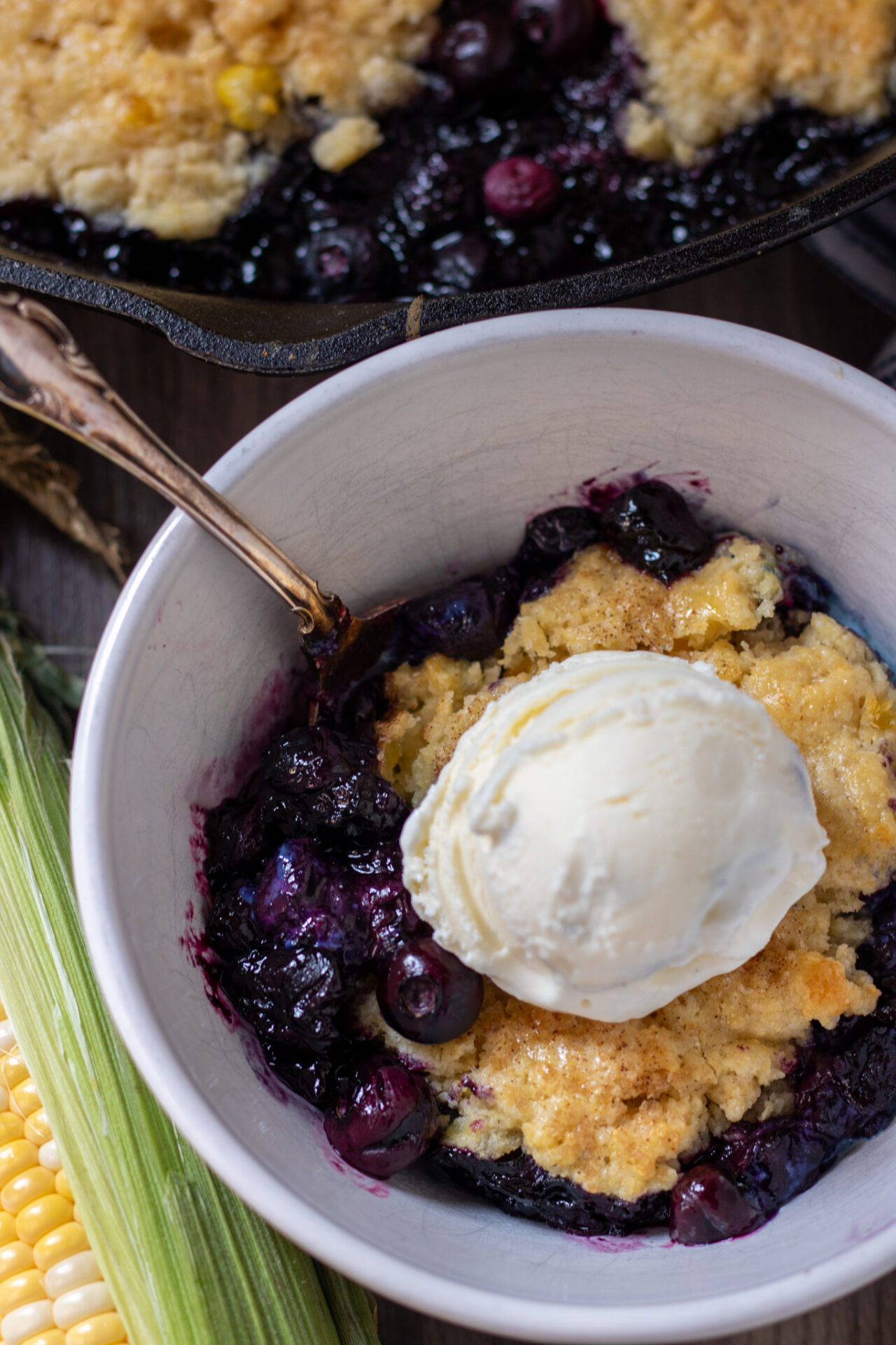 A white bowl with blueberry cobbler.  It's got a spoon in the bowl and it's topped with a scoop of vanilla ice cream.  There's an ear of fresh corn next to the bowl.