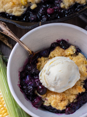 A white bowl with blueberry cobbler. It's got a spoon in the bowl and it's topped with a scoop of vanilla ice cream. There's an ear of fresh corn next to the bowl.