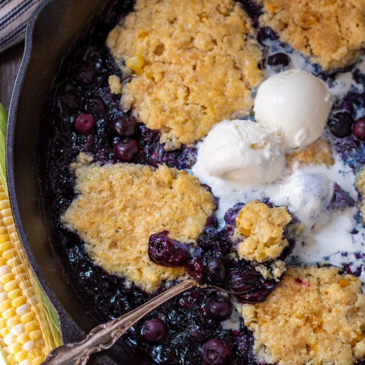 A cast iron skillet filled with blueberry cobbler. There's a spoon sticking into the cobbler with 3 scoops of slightly melted vanilla ice cream on top. There's a blue and white striped dish towel in the background and a fresh ear of corn.