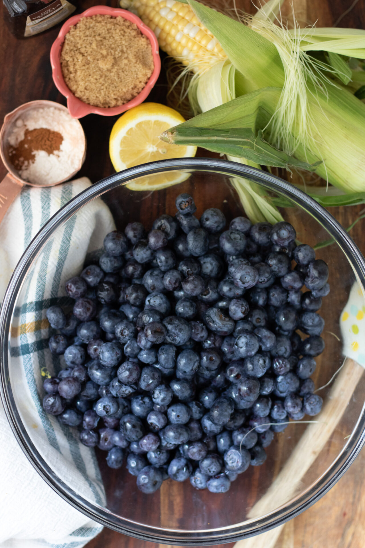 A large glass bowl filled with fresh blueberries. There's a small measuring cup with brown sugar, a half of a lemon, and an ear of corn in the background.