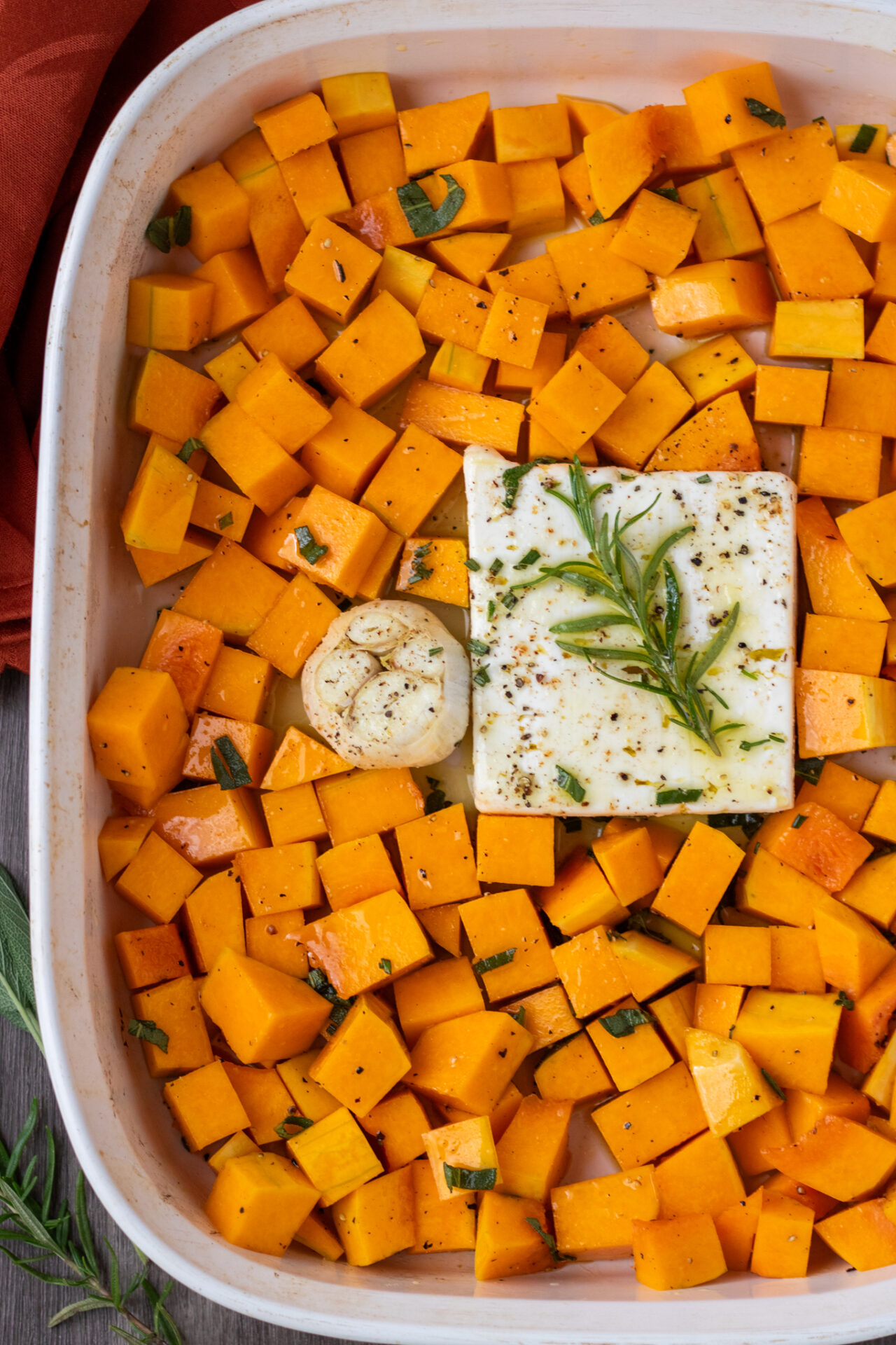 A large white baking dish filled with cubed butternut squash and a block of feta cheese in the middle. There's a whole head of garlic and a rosemary sprig.