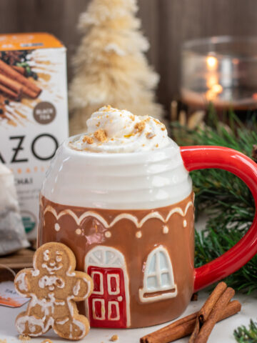 A gingerbread coffee mug filled with gingerbread chai tea. It's topped with whipped cream and cookie crumbles. There's a gingerbread cookie resting on the cup with two cinnamon sticks next to it. There's a candle in the background and white Christmas tree