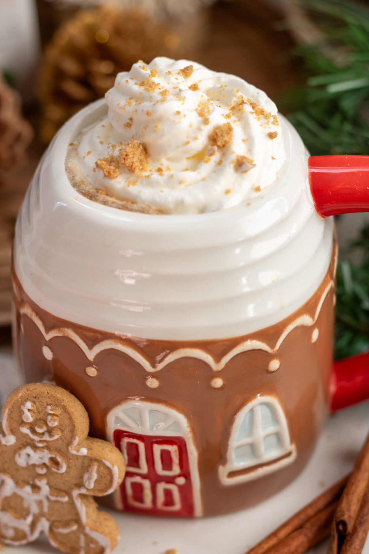 A gingerbread house coffee mug with gingerbread chai latte. It's topped with whipped cream and gingerbread cookie pieces. there's a gingerbread man cookie next to the cup and pinecones in the background